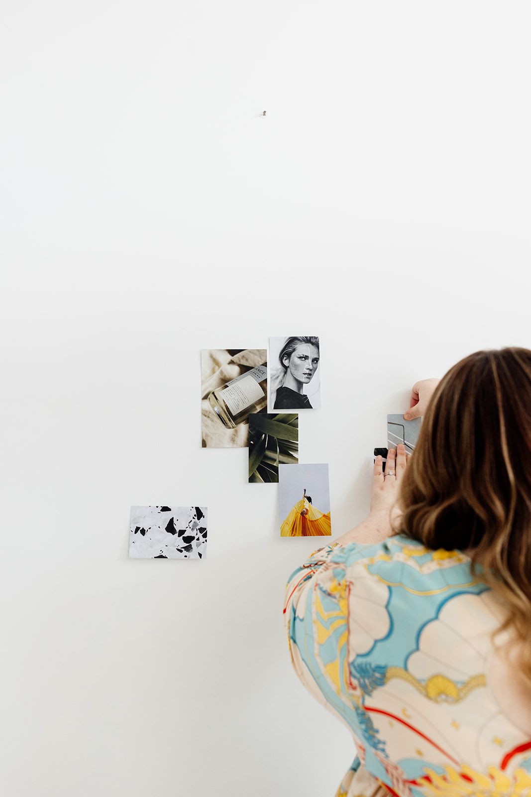 Image of a female creative entrepreneur creating a moodboard on a white wall with photographs, wearing a bright patterned top.