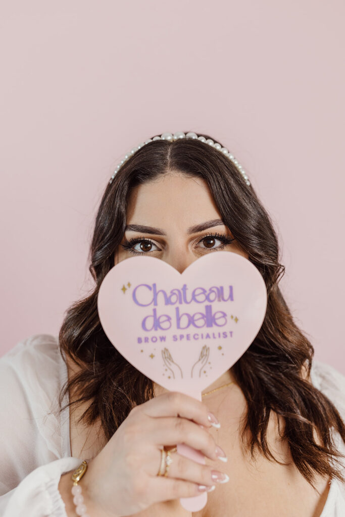 A close-up of a woman against a pink background. She wears a pearl headband and is holding a pink heart-shaped sign with the text "Chateau de belle" and "BROW SPECIALIST" written on it. The sign also features illustrations of hands.