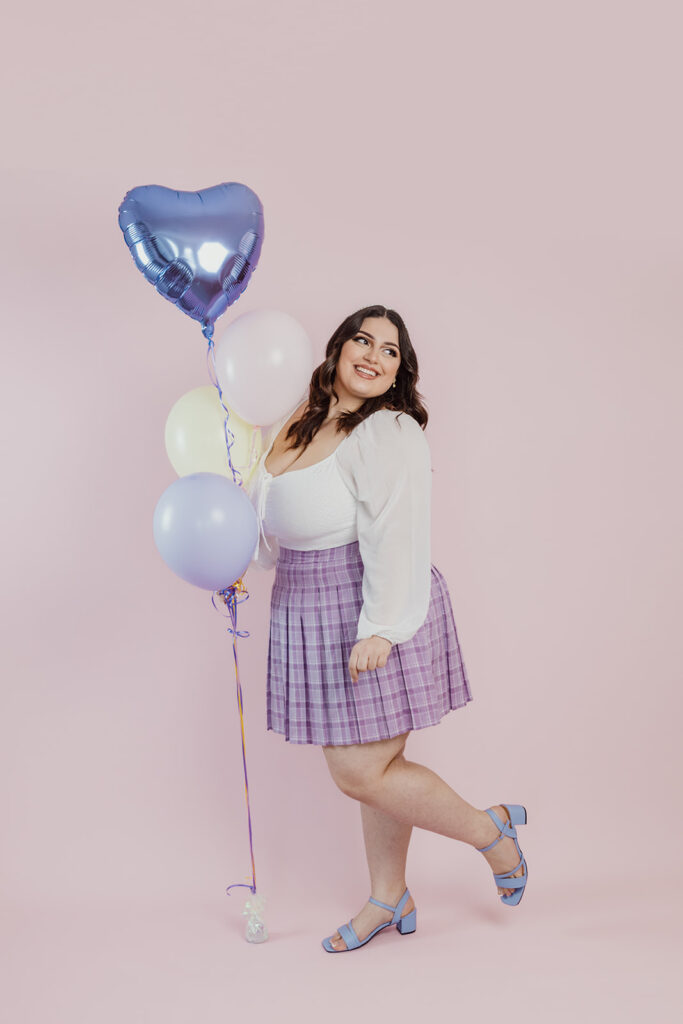 A female entrepreneur brow artist smiling and holding a bouquet of balloons, including a heart-shaped one, against a pink background. She wears a white blouse and a purple checkered skirt, paired with blue strapped heels.