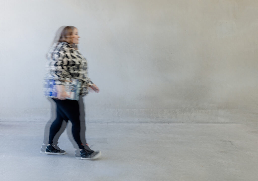 A blurred female branding designer walks against a light grey background, wearing a patterned jacket and dark leggings, symbolising dynamism and progress in a minimalist setting.
