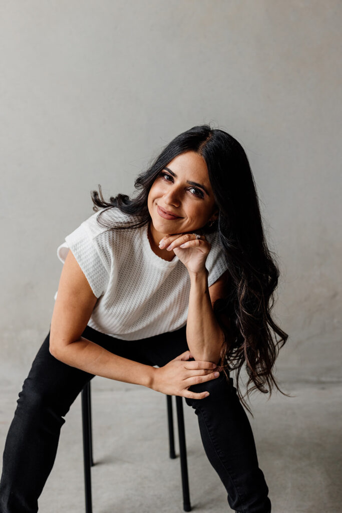 A female social media content creator with long dark hair, wearing a white top and black trousers, sits on a stool with a gentle smile, her hand resting on her chin against a neutral gray background.
