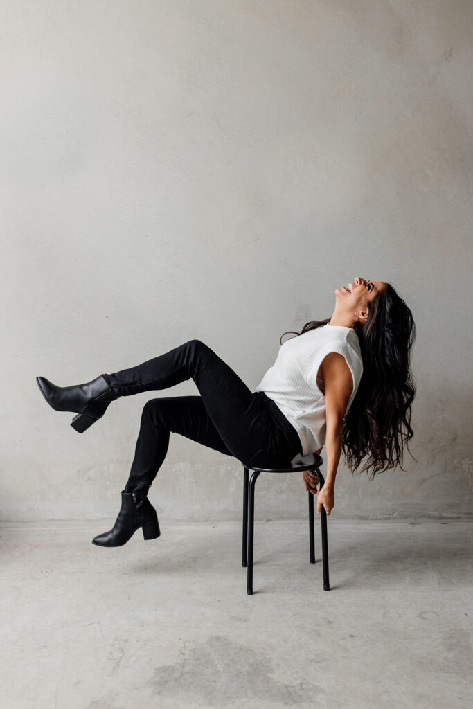 A female entrepreneur playfully leans back on the stool, laughing with her hair cascading down and legs elevated, showing black boots against a simple gray backdrop.